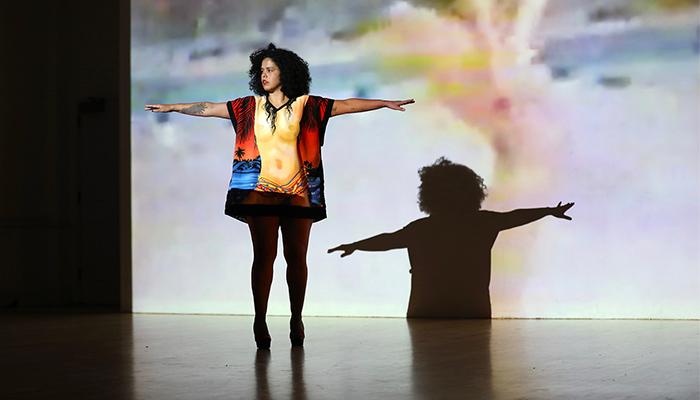 Awilda Rodríguez Lora  stands on a stage with arms outstretched wearing a shirt with a nude female torso