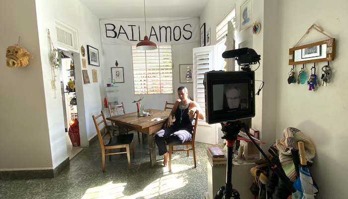 Choreographer Awilda Rodriguez Lora sits at a small, square dining table in her living and performance space La Rosario in Puerto Rico. A video camera recording her can be seen to the right in the forefront of the frame.