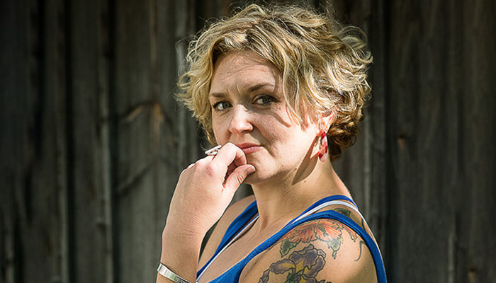 A photo of Abby Zbikowski in front of a darker background. Abby's left hand touches their chin. Abby has short blond/brown hair.