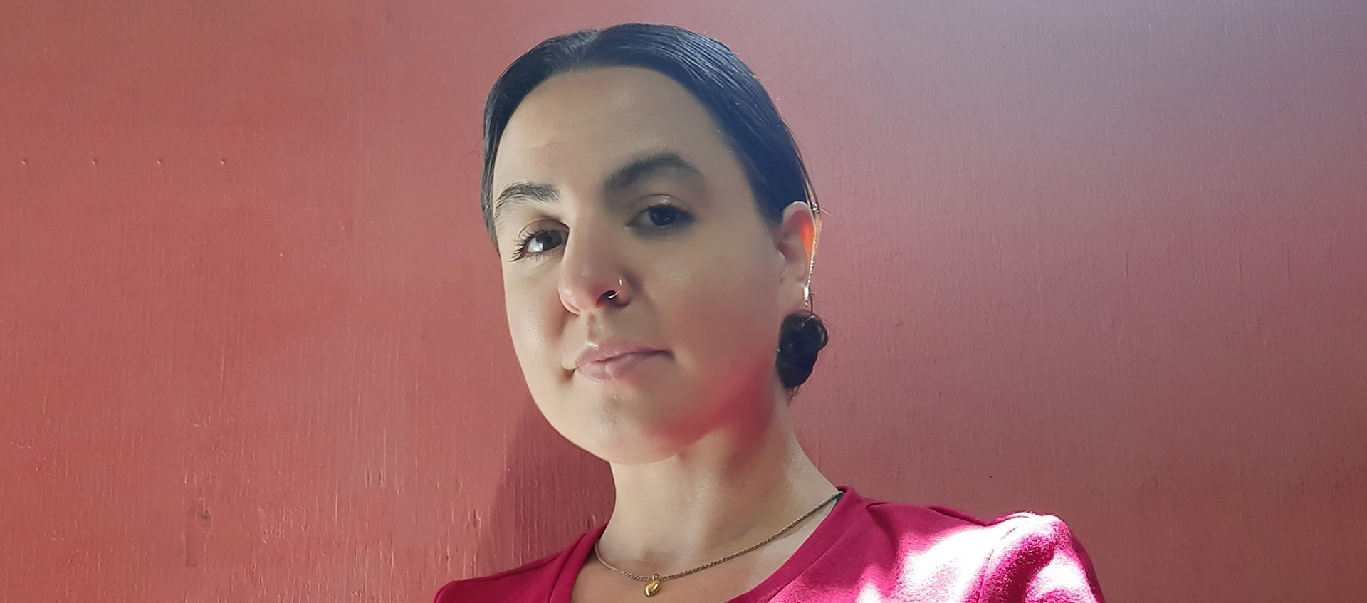 A photo of Tala Kanani in front of a reddish background. Tala has black hair and is wearing a red shirt, a gold necklace, and a nose ring.