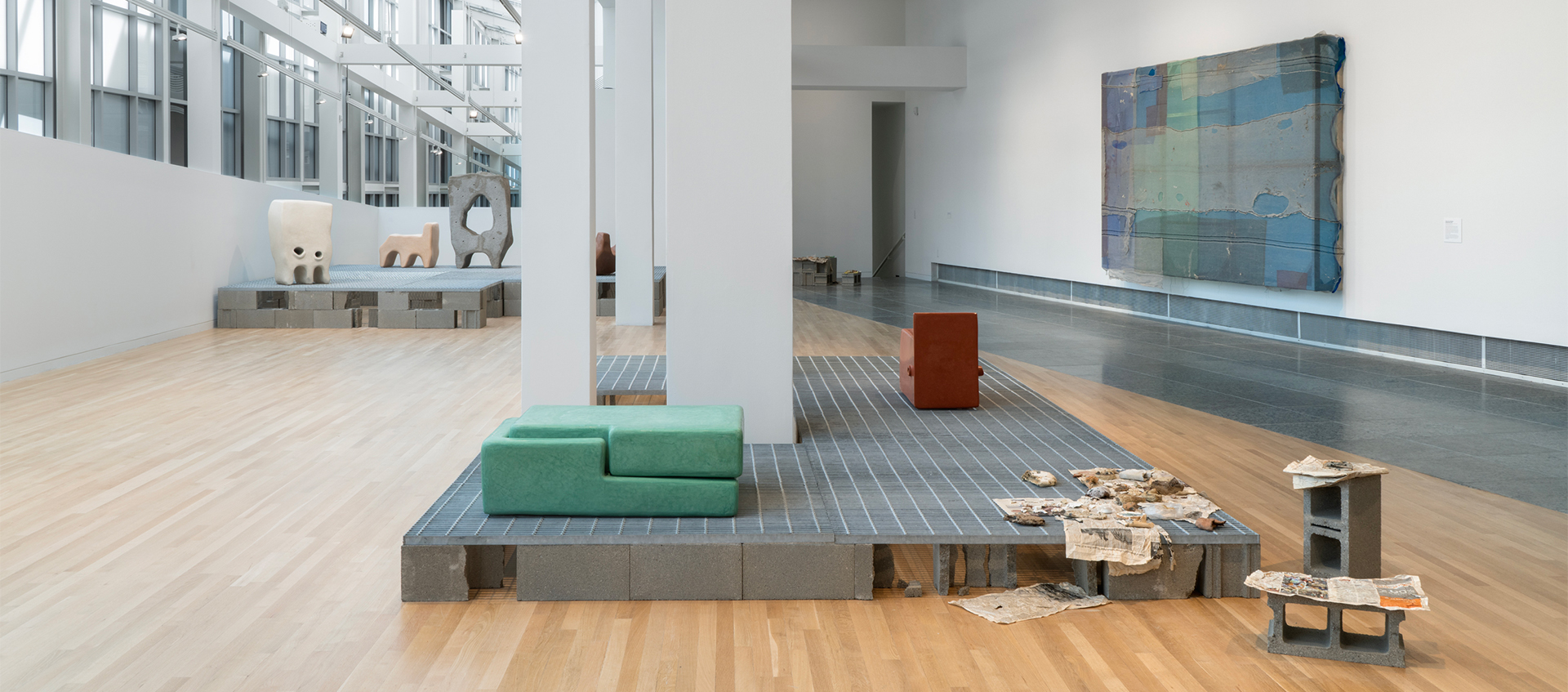 Installation of ceramic sculptures, including bread and a green double rectangle on metal grates on cinderblocks. Architecture is visible.