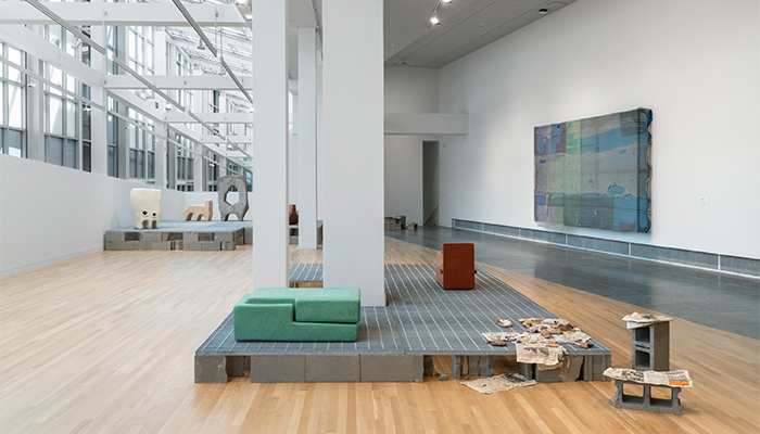 Installation of ceramic sculptures, including bread and a green double rectangle on metal grates on cinderblocks. Architecture is visible.