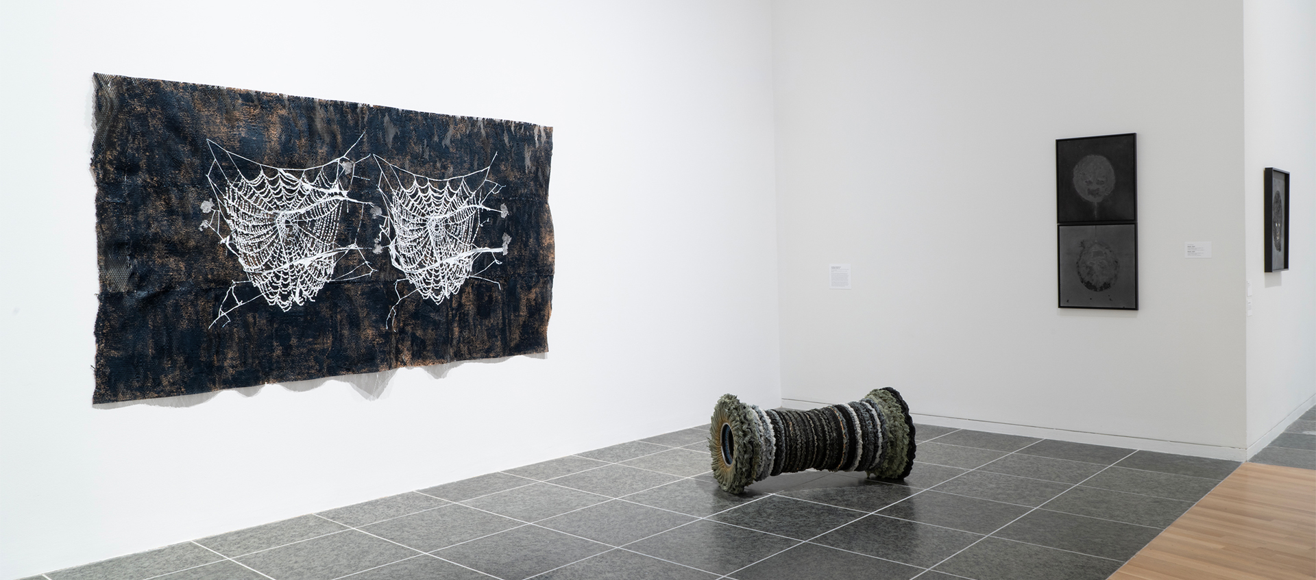 A polishing-wheel sculpture rests on the floor. On the two walls are a rectangular work with two spider webs and two stacked works. 