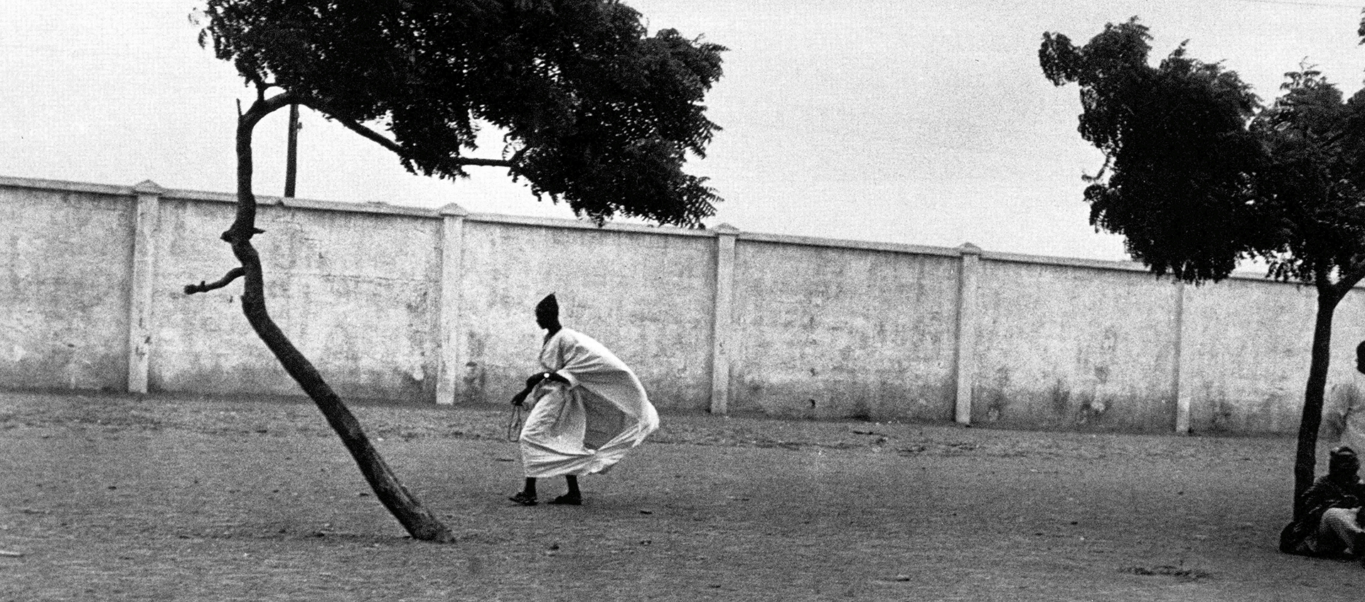 Panoramic photo of a Black person in a billowing white tunic walking next to a long, tall white wall. Two trees in the foreground frame the figure.