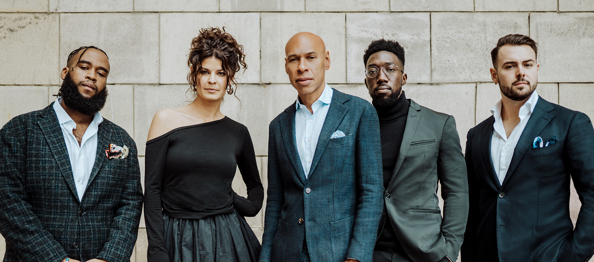 Four men and a woman stand against a stone wall, wearing suits and a dress. Joshua Redman stands in the center.