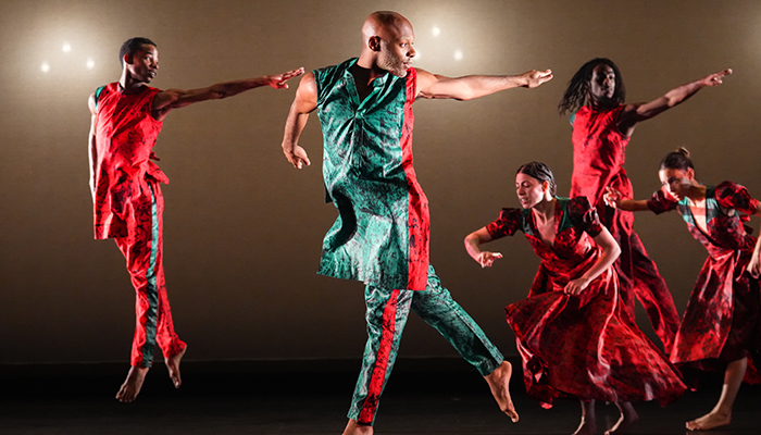 A group of dancers wearing red and blue costumes point to the right.