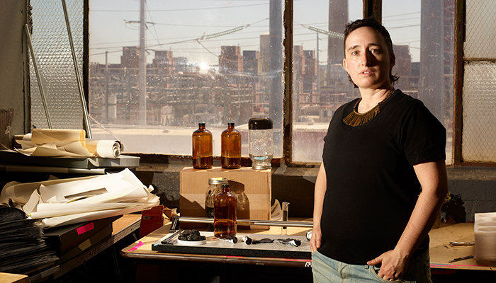 Jonas N.T. Becker stands in an industrial studio with large windows behind them. Jonas has a light skin tone, short dark hair, and wears a dark shirt.