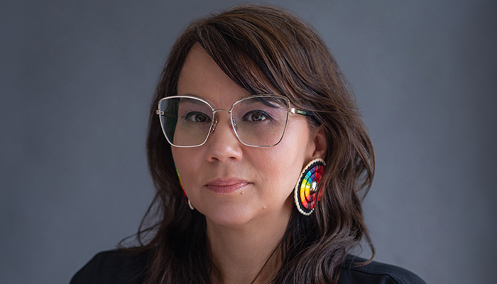 Portrait of Tanya Lukin Linklater. She has long brown hair, a medium skin tone, and wears geometric silver glasses, beaded earrings, and a dark shirt.