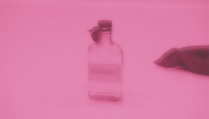 A blurry, pink-tinged image of a clear, lidded sloped shoulder, flattened glass bottle labeled with the word susteno.