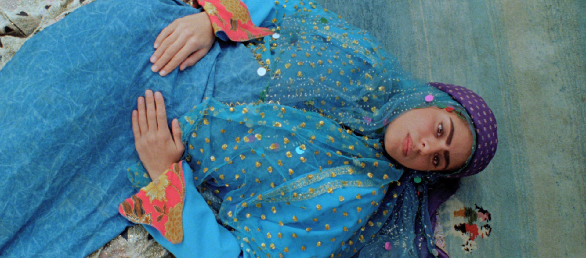 A woman wearing bright blue robes lays on a rug with her hands on her stomach.