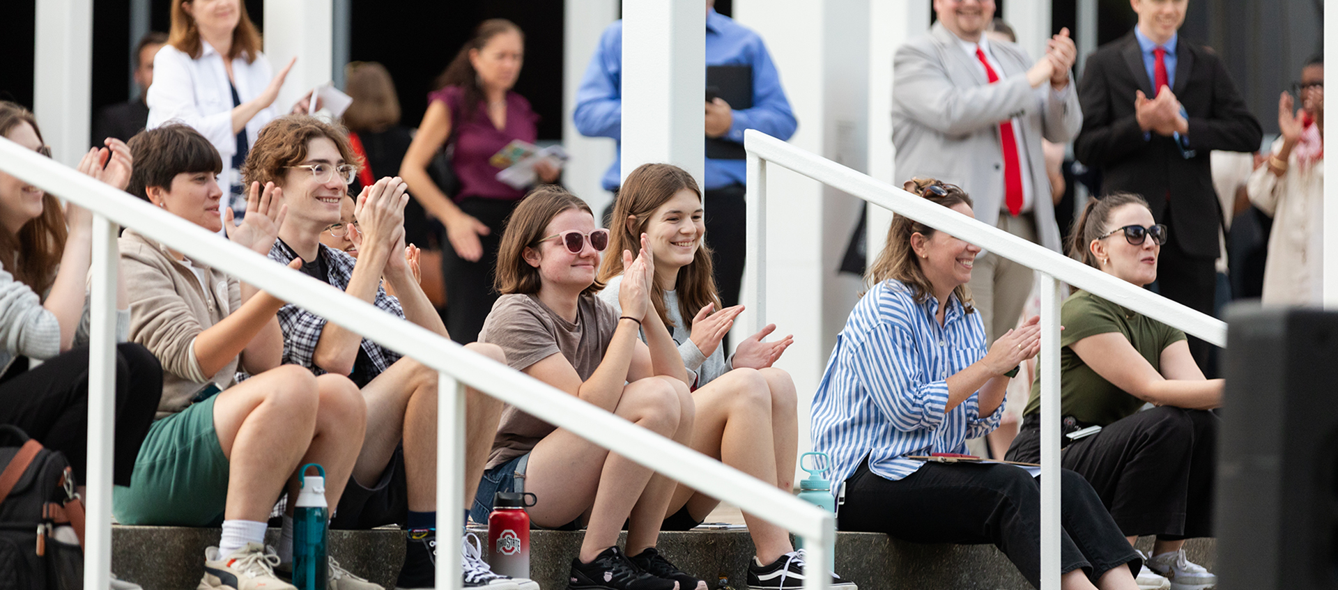 Several people smile and applaud as they sit on steps or stand in the background and look to the right at a performance outside the image frame.