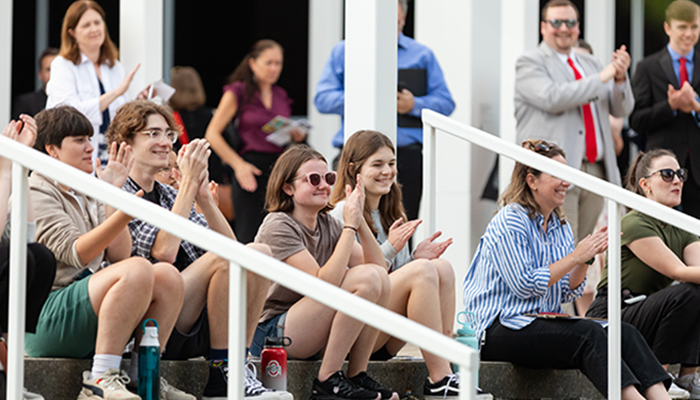 Several people smile and applaud as they sit on steps or stand in the background and look to the right at a performance outside the image frame.