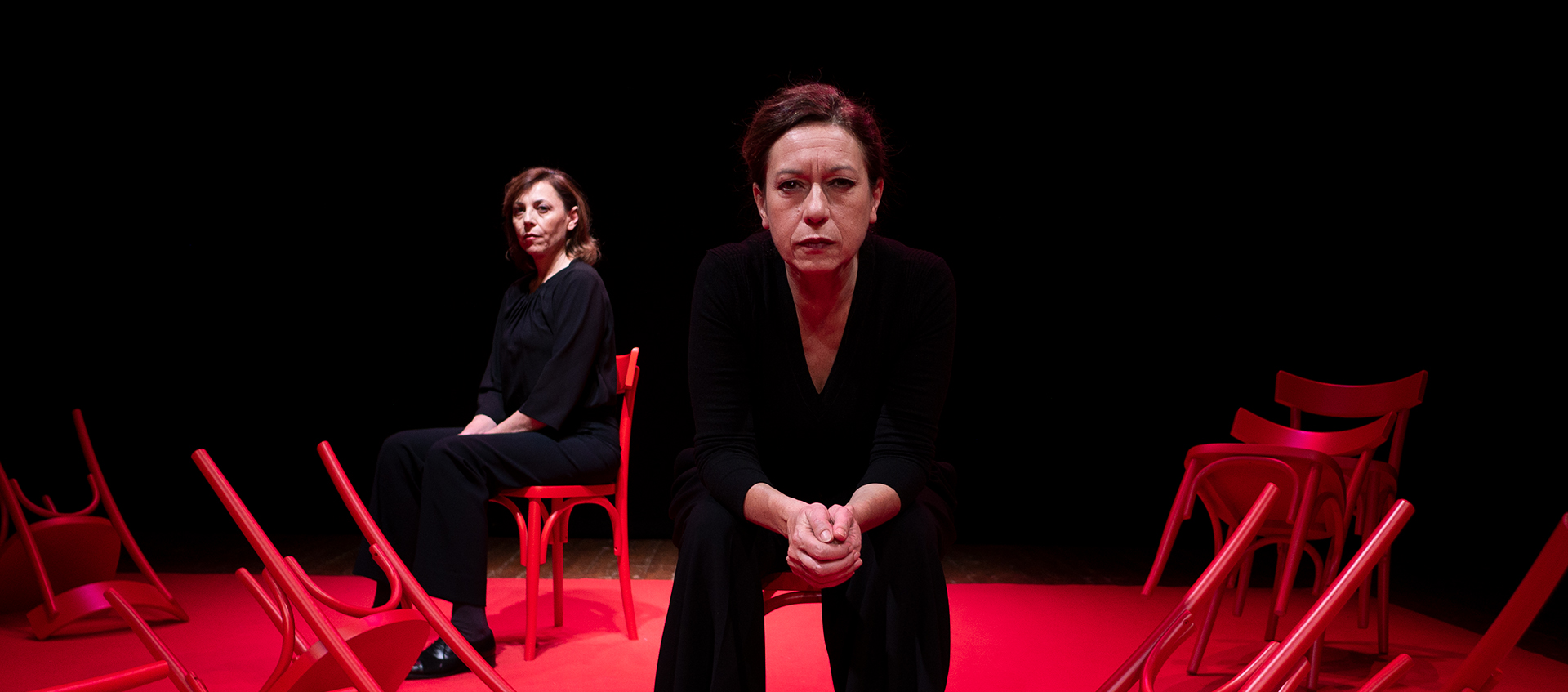 a woman sits on a red stage with a black background on a chair wearing a suit with another woman sitting behind her.