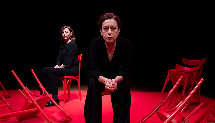 a woman sits on a red stage with a black background on a chair wearing a suit with another woman sitting behind her.