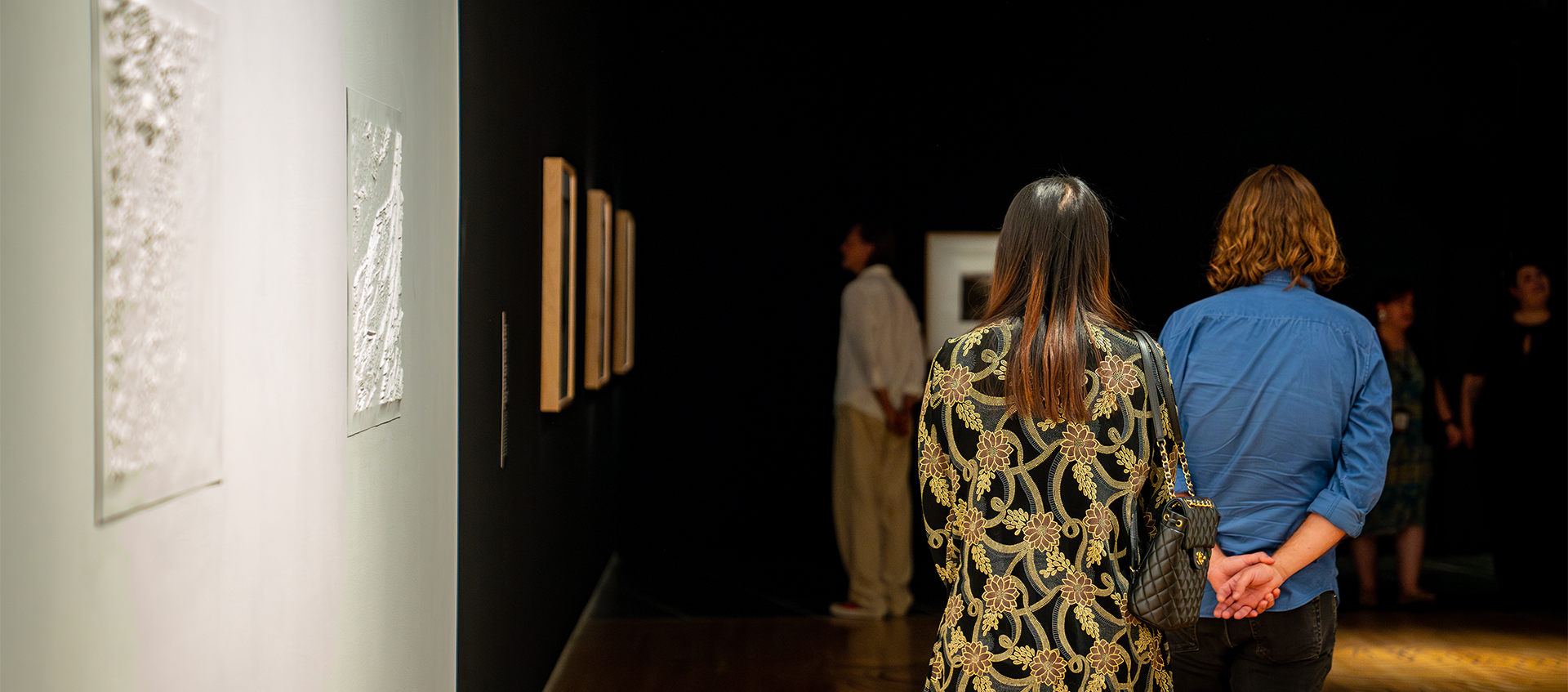 Two people walk out of a white gallery space displaying white topographical maps and into a dark gallery with framed photographs.