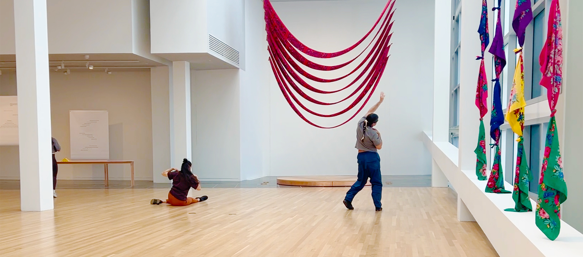 Three dancers face away from the camera and gesture with their arms in a gallery. Two stand at left and right, while the third is seated left of center. 