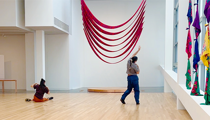 Three dancers face away from the camera and gesture with their arms in a gallery. Two stand at left and right, while the third is seated left of center. 