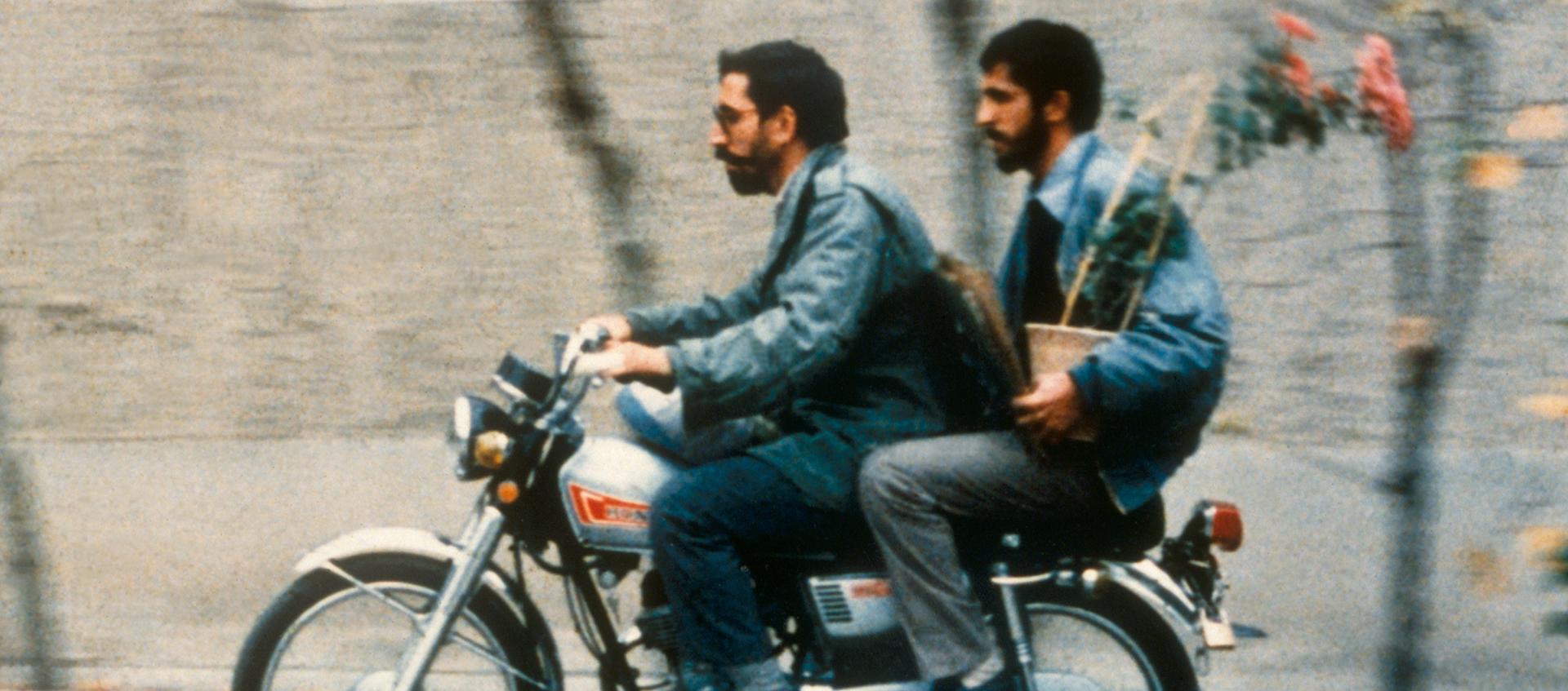 Two bearded men are riding on a motorcycle together wearing blue jackets. The man in front is wearing sunglasses.  