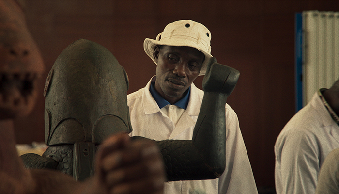 A Black man wearing a lab coat and bucket hat is looking at a statue.