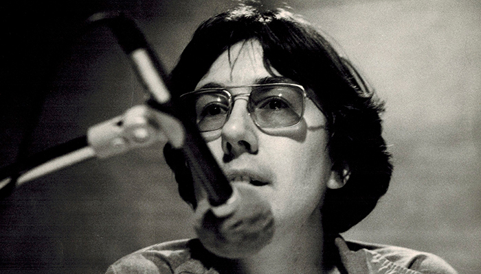 Julia Reichert in the mid 1970s, she has short hair, glasses and is in front of a microphone.