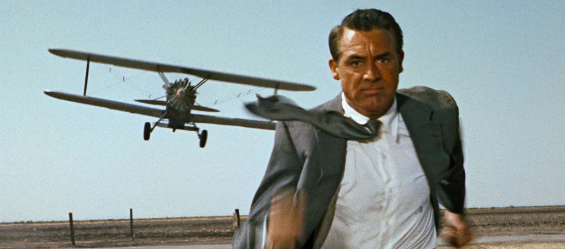 A man is running toward the camera in a barren field. He is wearing a gray suit. A small plane can be seen over his right shoulder flying toward him.