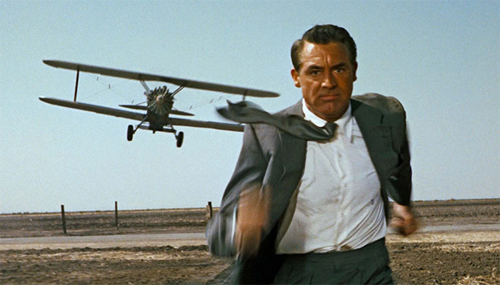 A man is running toward the camera in a barren field. He is wearing a gray suit. A small plane can be seen over his right shoulder flying toward him.