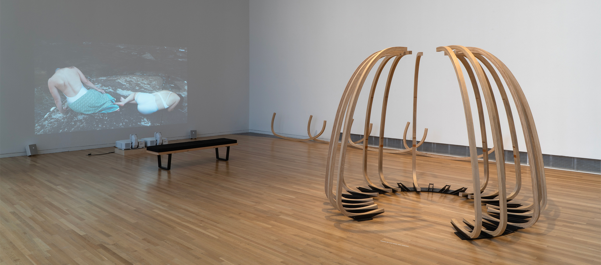 A tall, semicircular sculpture of fifteen curved wooden ribs on a metal base sits near a video projection depicting two figures reclining on a rock. 