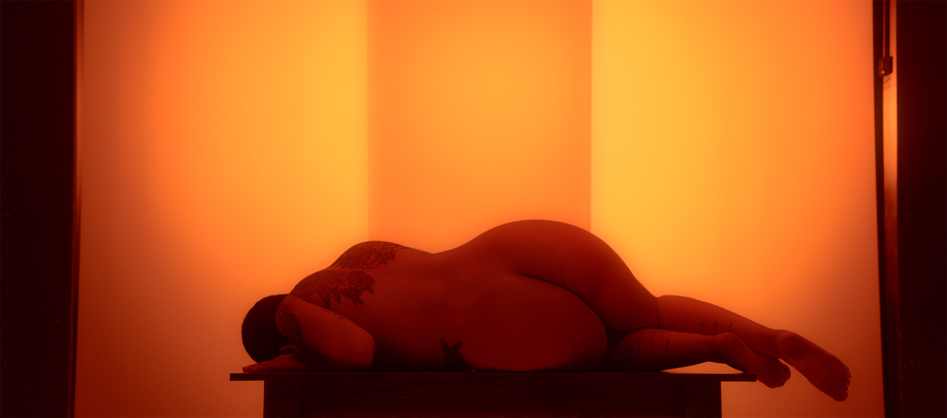 The artist lies naked on a table against a backlit amber background. She is on her side, facing away from the camera, with her head turned downward.