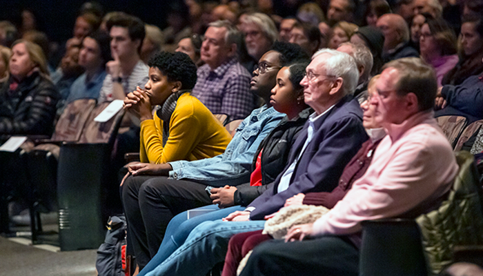 A closeup of a crowded theater where an audience looks attentively toward the stage, which is outside of the left frame of the photograph.