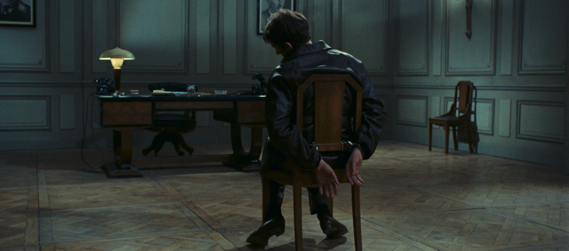 A man is tied to a chair in the middle of a large room. There is a desk in front of him and two photos of military officers on the otherwise empty walls.