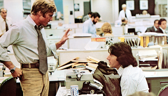 Two men are talking in a busy newsroom, one of them is standing and gesturing while the other sits. 