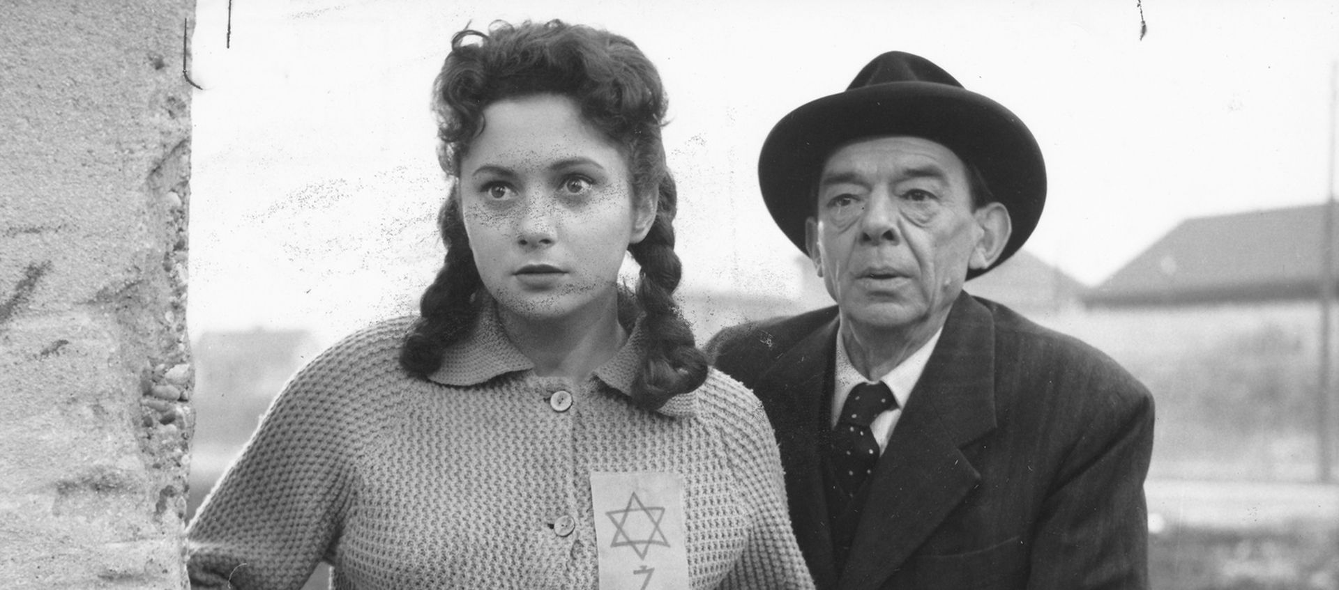 A young woman wearing a Star of David patch and an older man in a suit are standing next to each other looking around the corner of a building. 