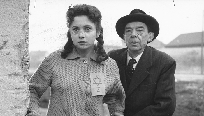 A young woman wearing a Star of David patch and an older man in a suit are standing next to each other looking around the corner of a building. 