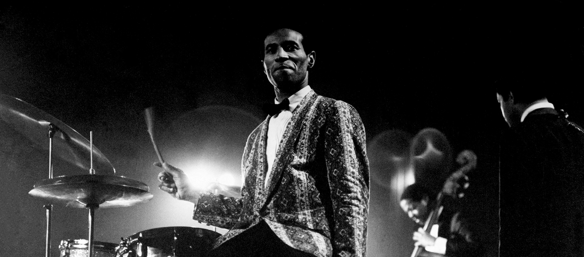 A Black man wearing a pattered jacket and bow tie sits at a drum set, his right hand is in motion. 