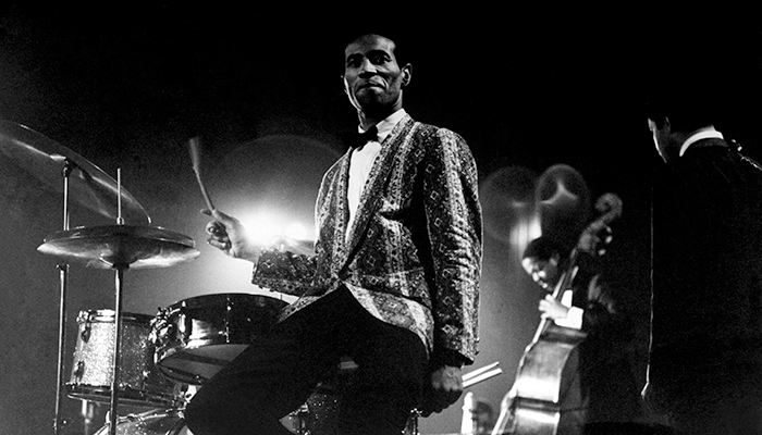 A Black man wearing a pattered jacket and bow tie sits at a drum set, his right hand is in motion. 