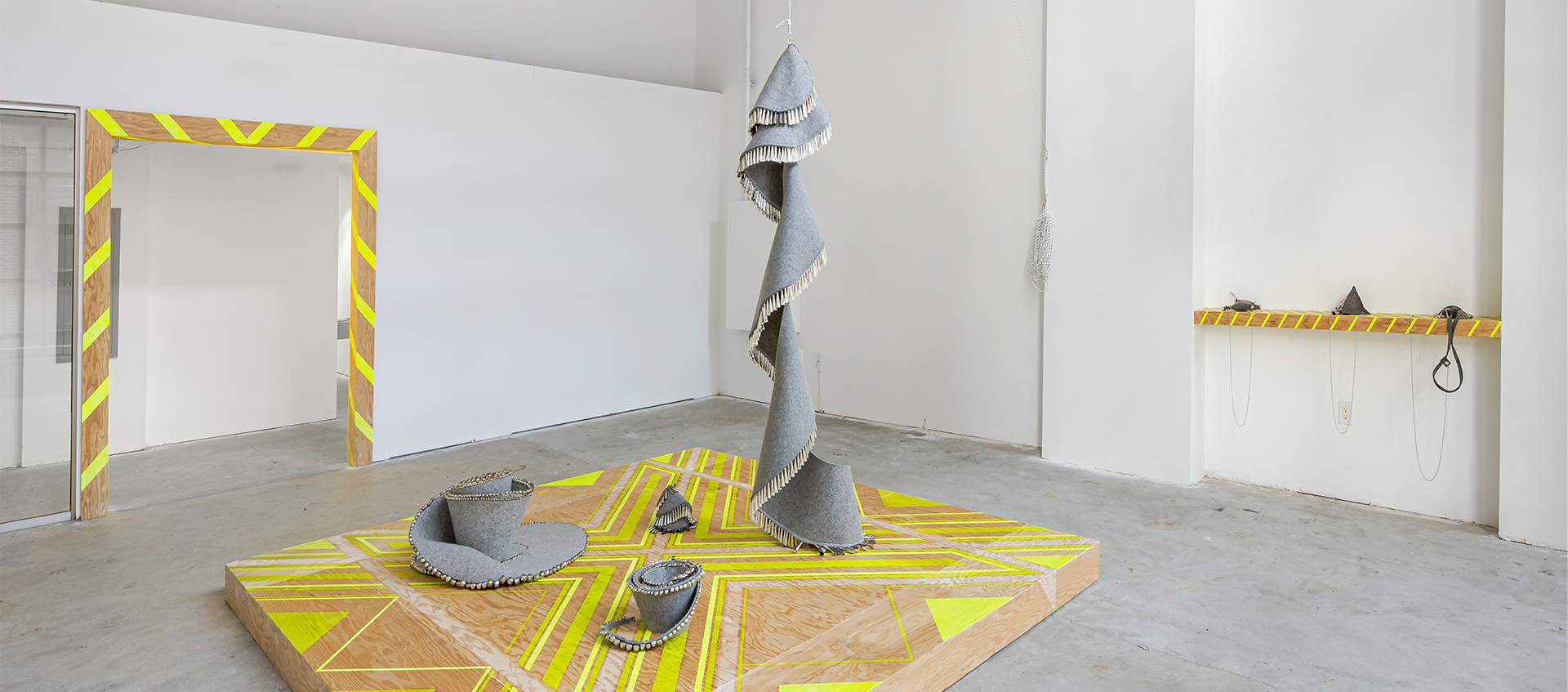 A wooden plinth with a yellow geometric pattern supports three gray felt and jingle sculptures; a fourth hangs and spirals down onto the platform.