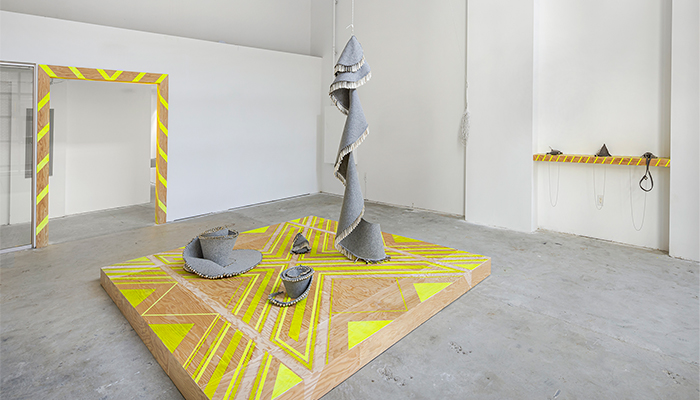 A wooden plinth with a yellow geometric pattern supports three gray felt and jingle sculptures; a fourth hangs and spirals down onto the platform.