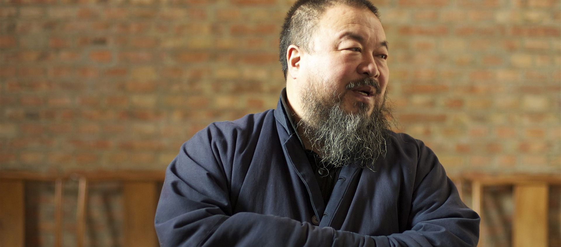 Photo of Ai Weiwei, a man with a beard wearing a blue jacket with his arms folded standing in front of a brick wall.