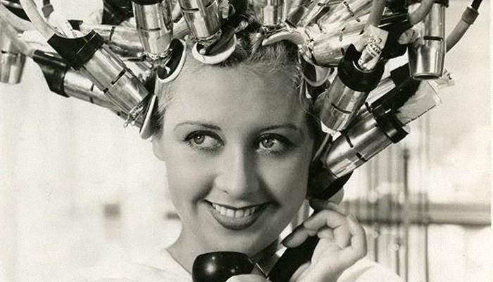 Black and white image of a smiling woman with her hair up in a large hair curling machine.