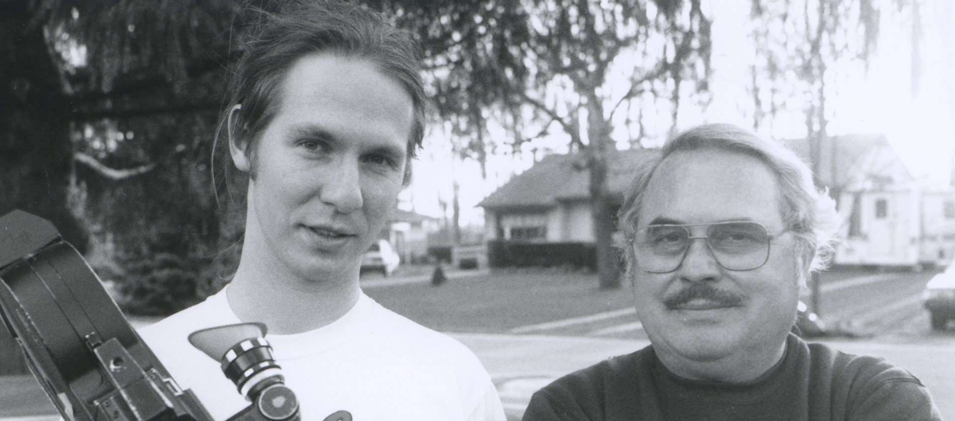 Black and white image of a young man holding a film camera standing next to an older man whose arms are crossed.