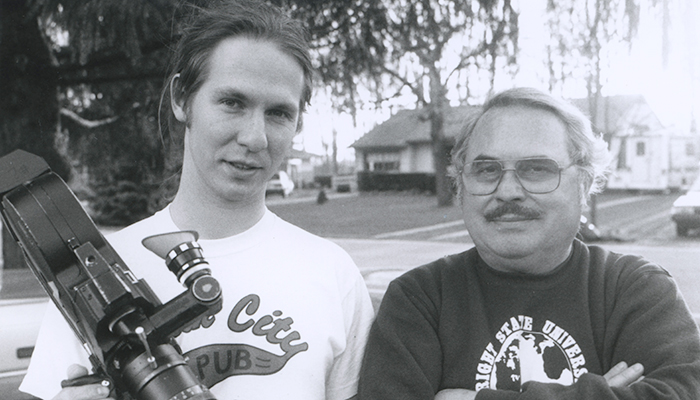 Black and white image of a young man holding a film camera standing next to an older man whose arms are crossed.