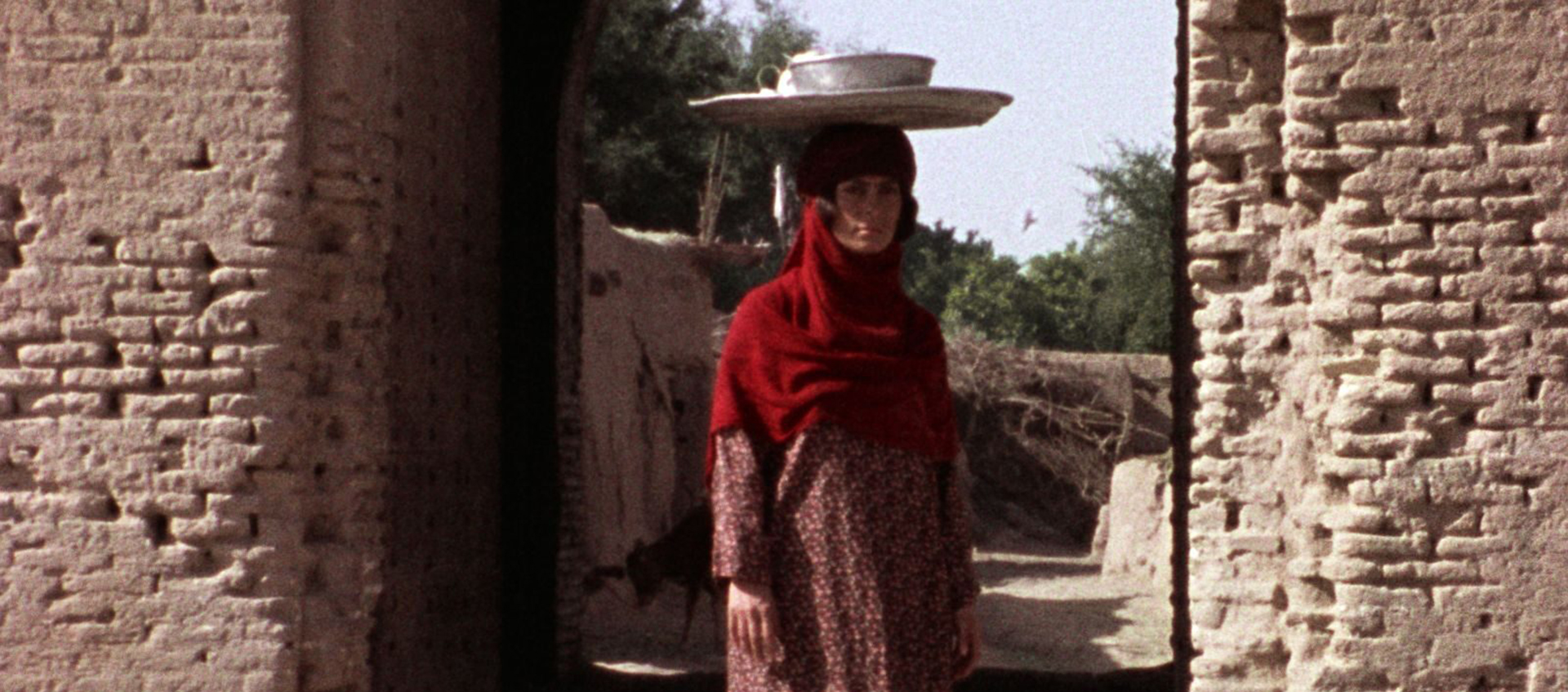 A woman is standing under the arch of a very old looking building. She is balancing a tray and bowl on her head.