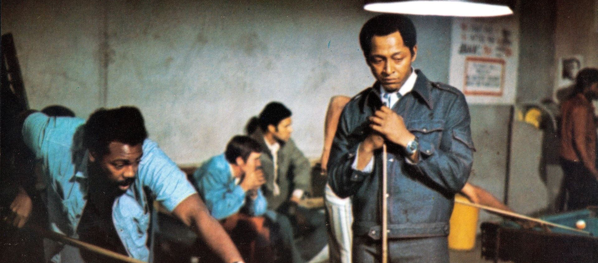 Two Black men playing pool in a pool hall. One man is bent over shooting, the other is standing nearby watching.