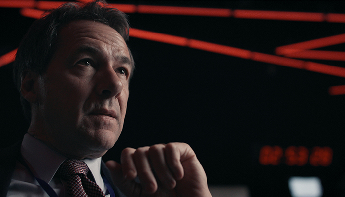 A closeup of a white, middle-aged man in a suit looking offscreen. He is in a dark situation room with dramatic red lines of lights on the ceiling.