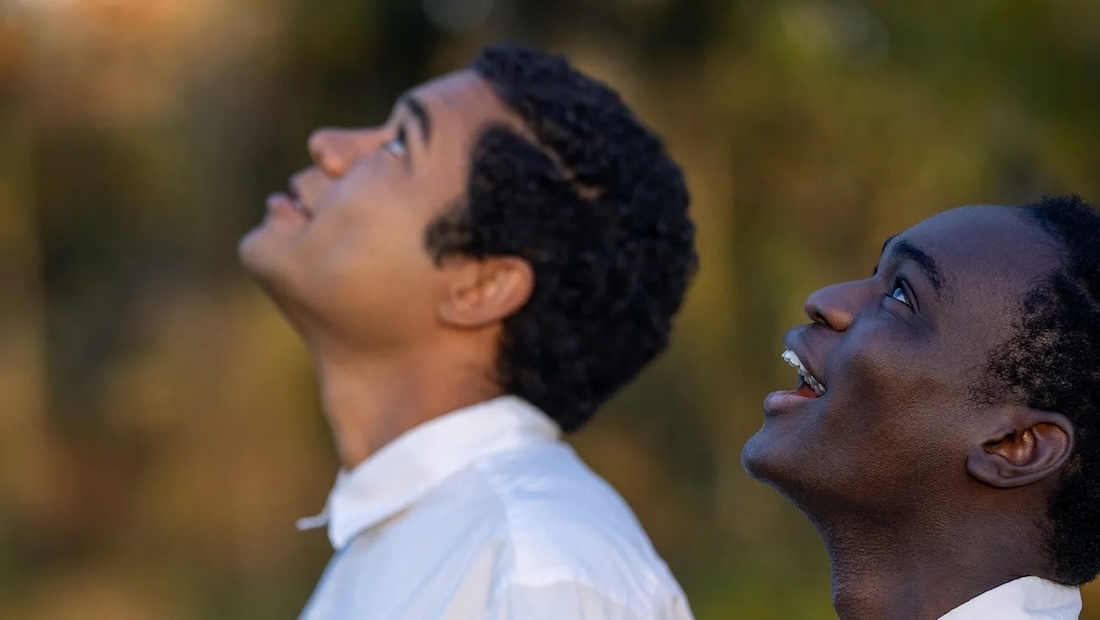 Two young Black men, seen in profile, stand outside and look upward, smiling.