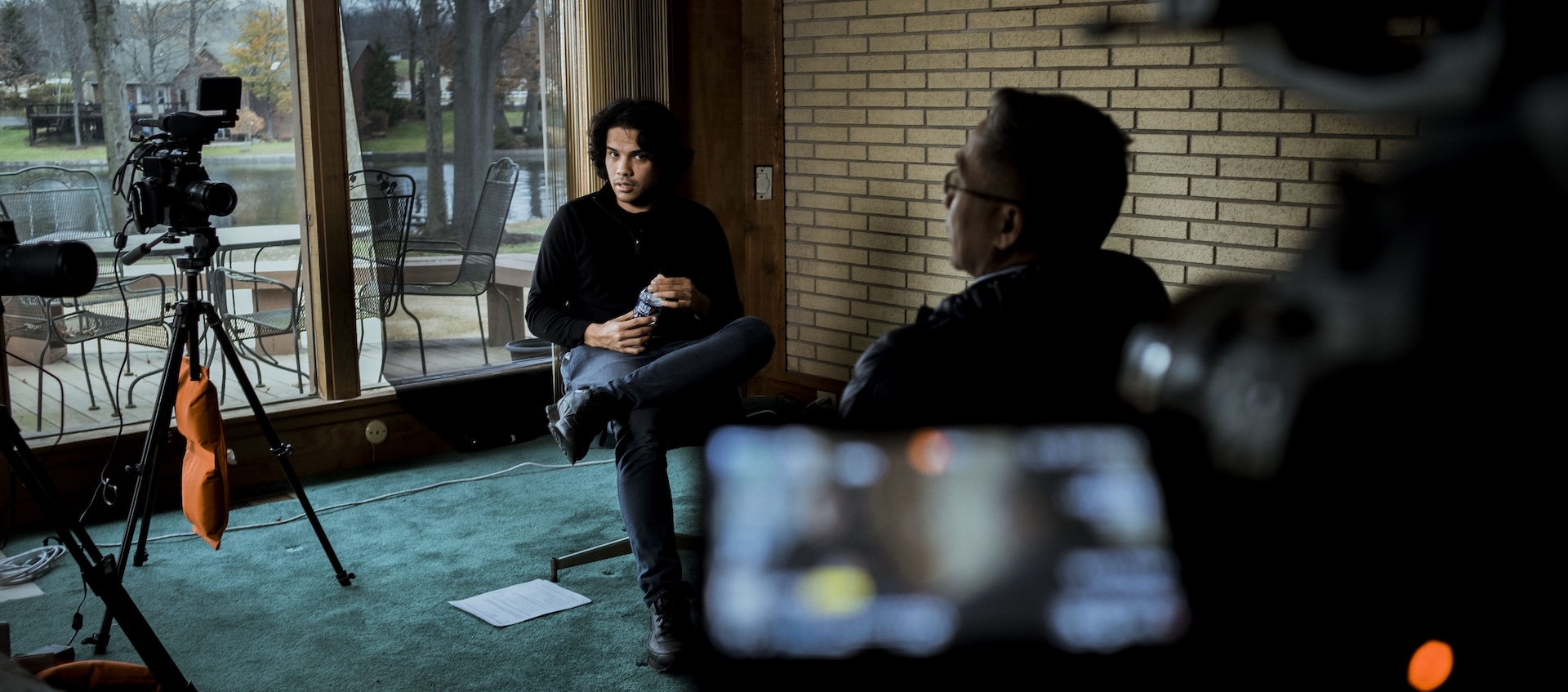 An interview between a young man and an older man is recorded by video cameras inside a living room.