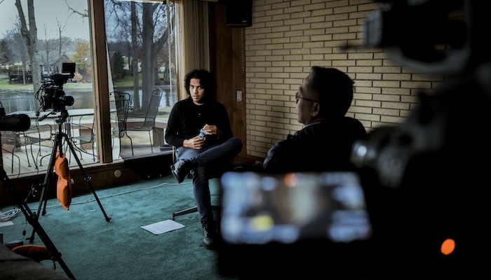 An interview between a young man and an older man is recorded by video cameras inside a living room.