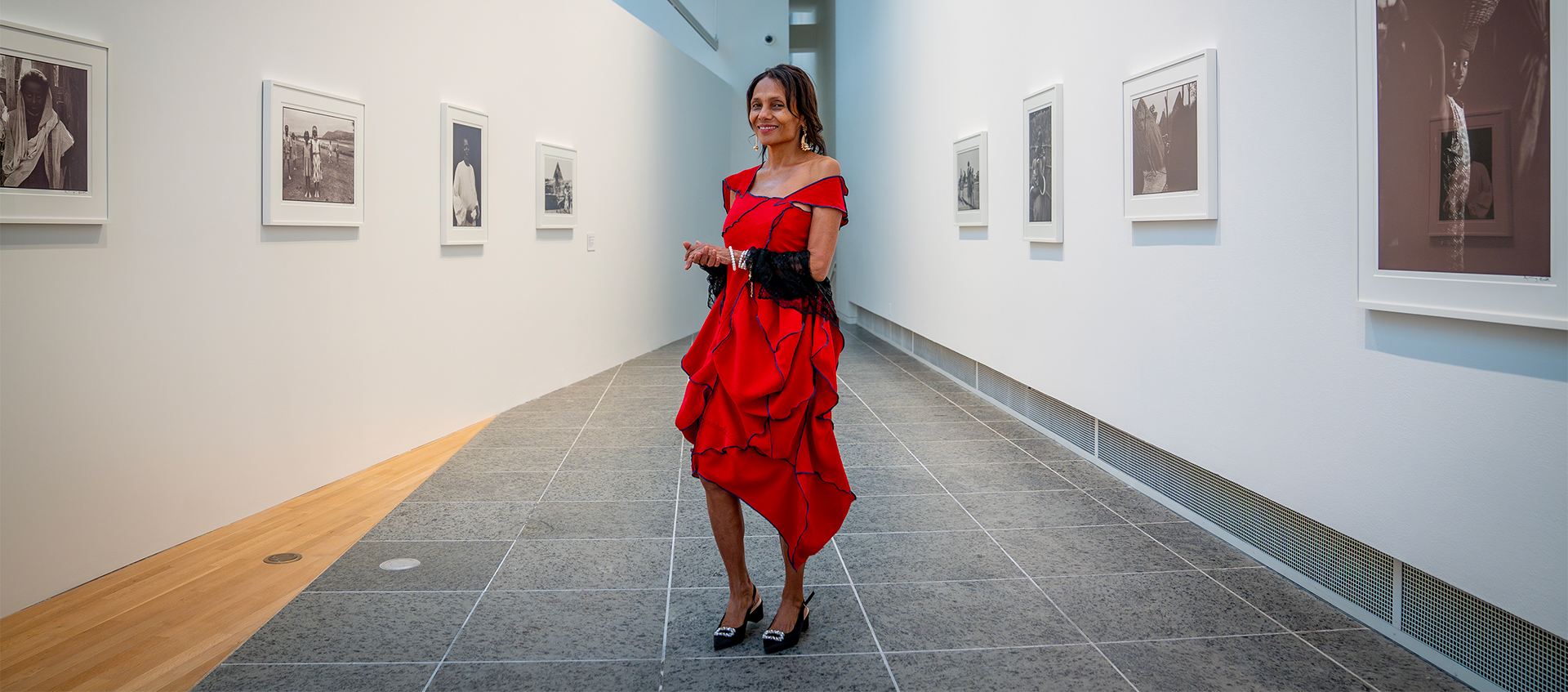 Ming Smith wears a red dress with black stitching and stands, smiling in a long narrow gallery featuring her photographs on the walls. 