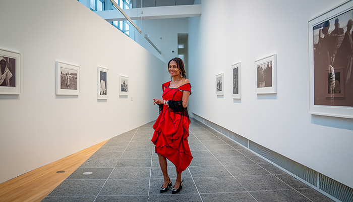 Ming Smith wears a red dress with black stitching and stands, smiling in a long narrow gallery featuring her photographs on the walls. 