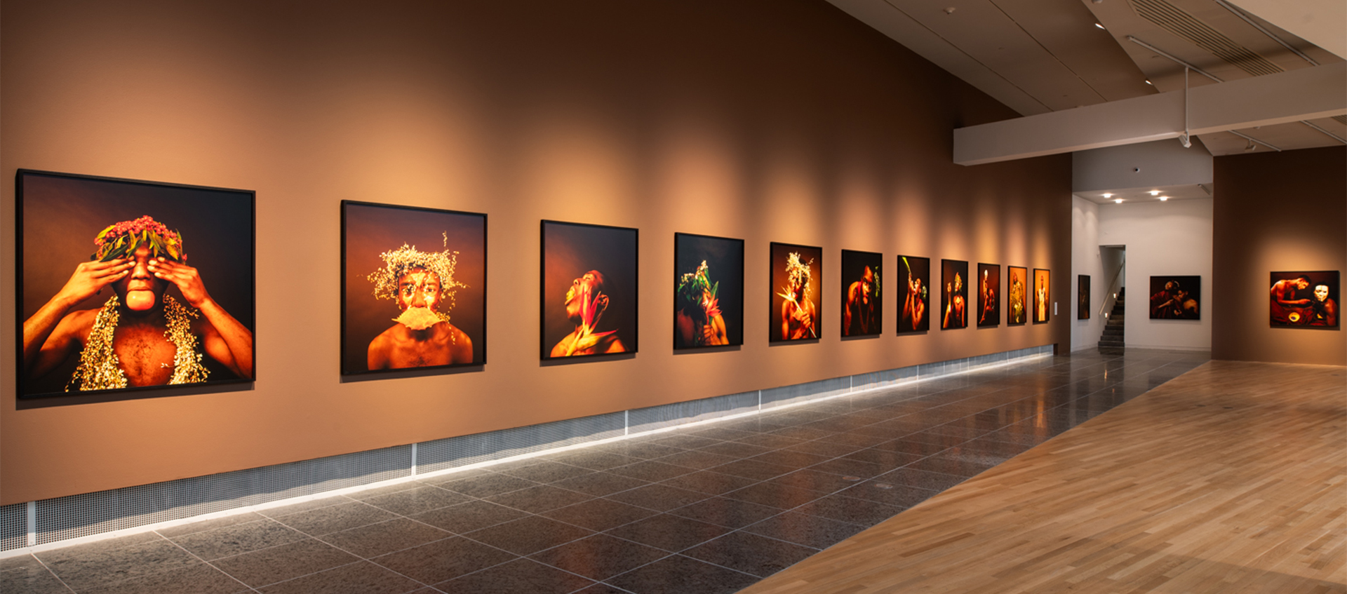 View of 14 large-scale color photographs on a brown gallery wall. The images show young Black men wearing or interacting with plants, fruit, or shells.